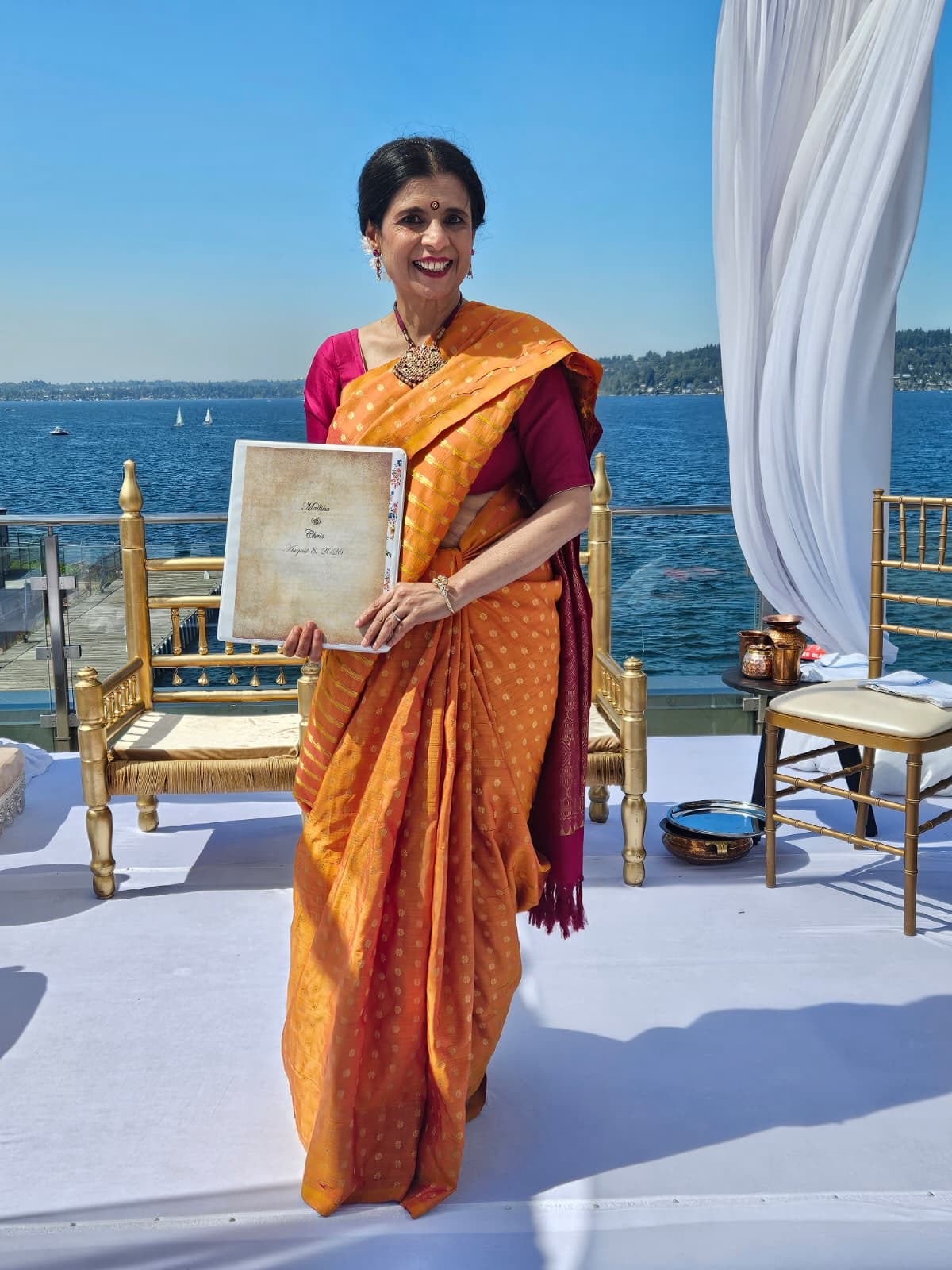 Dr. Meenakshi Rishi in an orange sari holding the couple's ceremony program, with the lake behind her