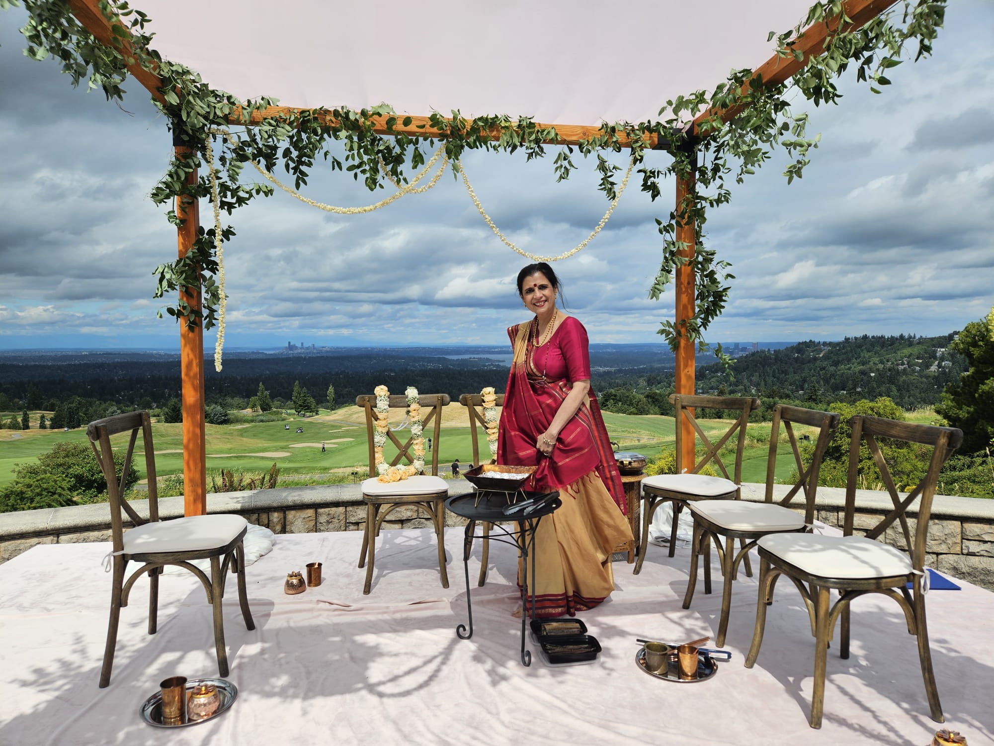 Dr. Meenakshi Rishi beside the havan setup at an outdoor wedding pavilion overlooking the hills and city